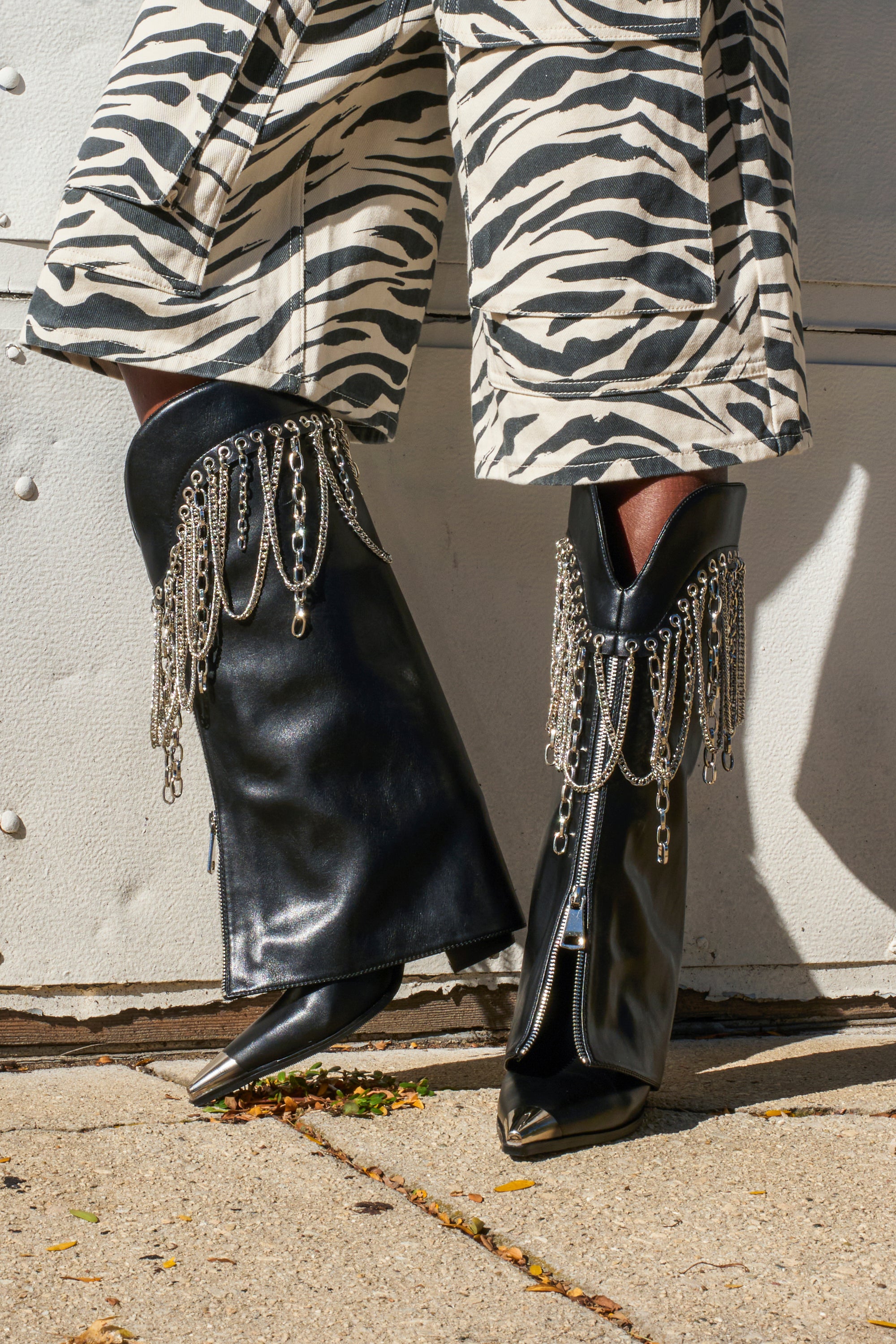 A person wearing zebra-print pants and AZALEA WANG SINISTER BLACK WESTERN CHUNKY BOOT with silver chains stands on a sidewalk.