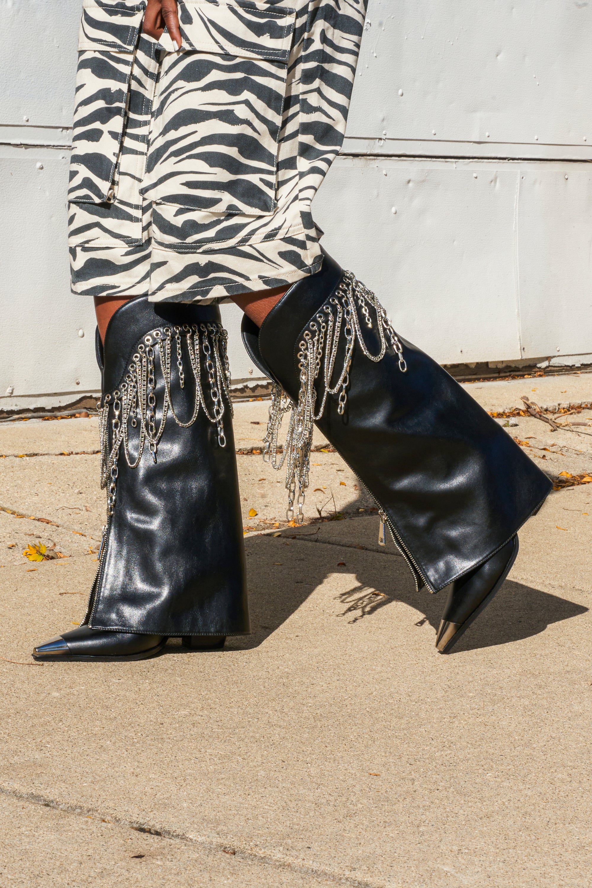 A person in a zebra-print skirt walks on a sunny sidewalk, wearing AZALEA WANG SINISTER BLACK WESTERN CHUNKY BOOTS decorated with silver chains.