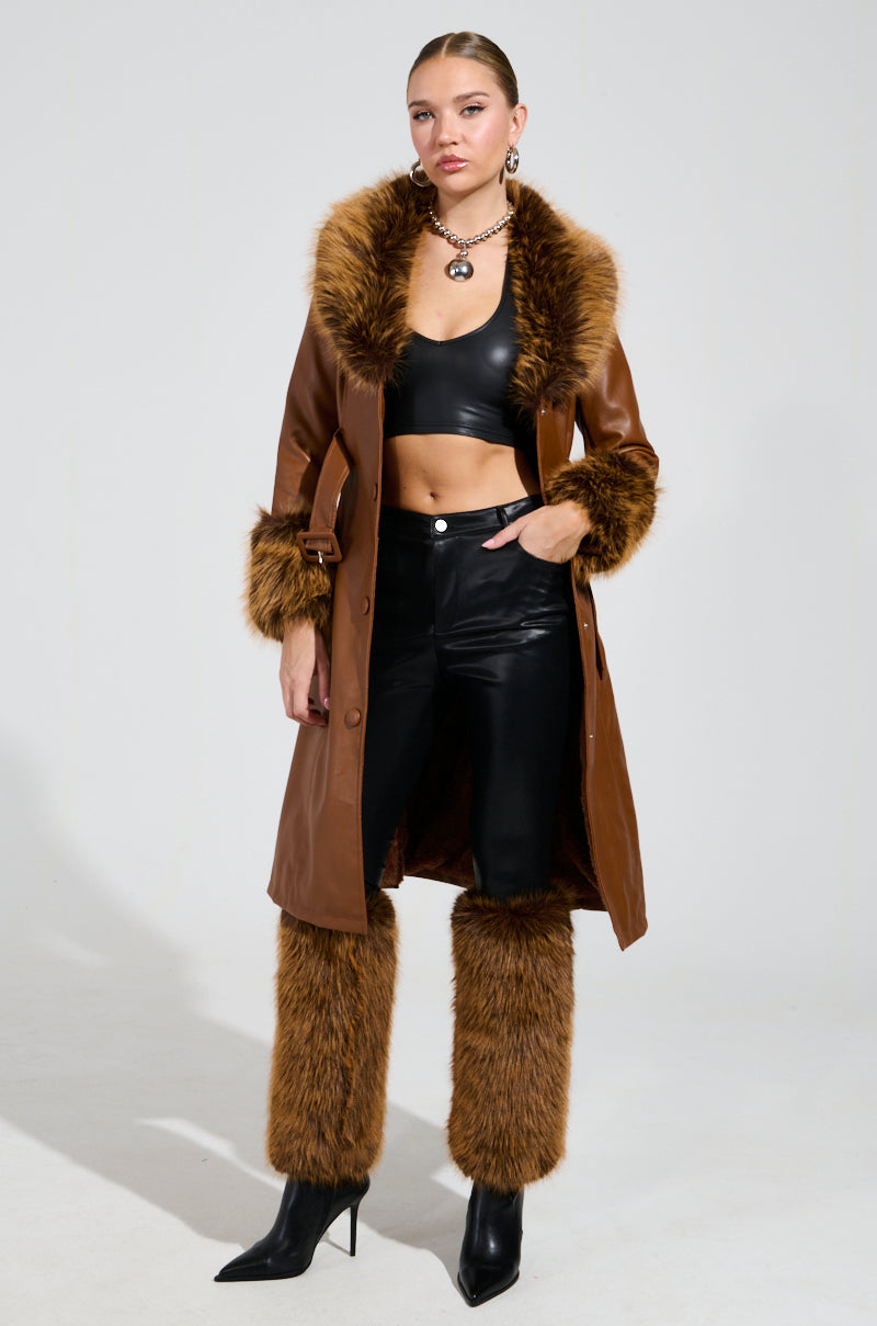 A person poses confidently in a stylish outfit, showcasing a long brown leather coat with faux fur trim. They wear the flattering black SIREN FUR TRIM FAUX LEATHER PANTS paired with a curve-hugging black crop top, faux fur leg warmers, and sleek heeled ankle boots. Accessories include a necklace and hoop earrings.
