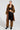 A person poses confidently in a stylish outfit, showcasing a long brown leather coat with faux fur trim. They wear the flattering black SIREN FUR TRIM FAUX LEATHER PANTS paired with a curve-hugging black crop top, faux fur leg warmers, and sleek heeled ankle boots. Accessories include a necklace and hoop earrings.