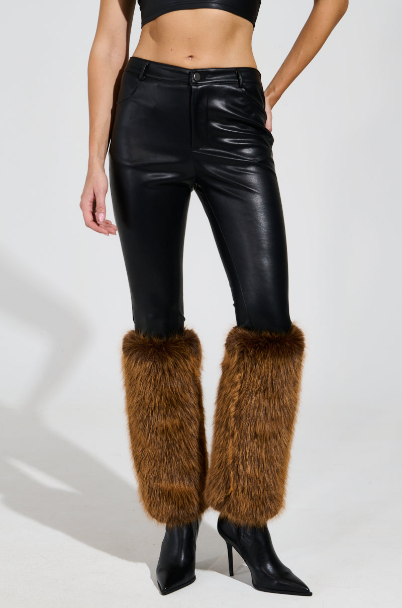 A person styled in the SIREN FUR TRIM FAUX LEATHER PANTS featuring brown faux fur accents at the ankles, paired with a black crop top and sleek black pointed boots, all set against a plain, light-colored backdrop.