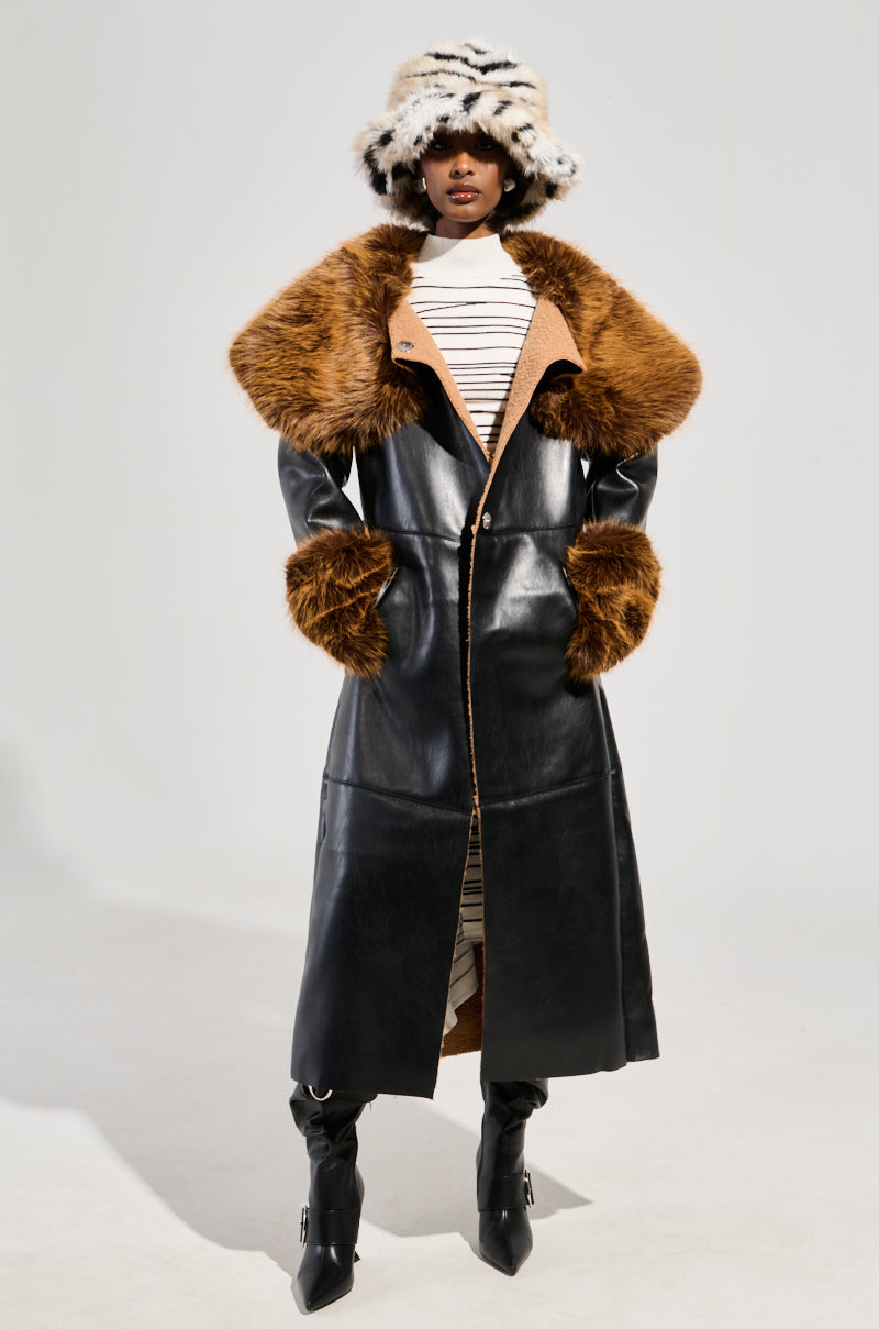 The individual is dressed in the SIREN FUR TRIM TRENCH COAT, a long black faux leather trench coat featuring prominent brown faux fur accents at the collar and cuffs. They complement this attire with a striped hat and boots, standing against a simple white background.