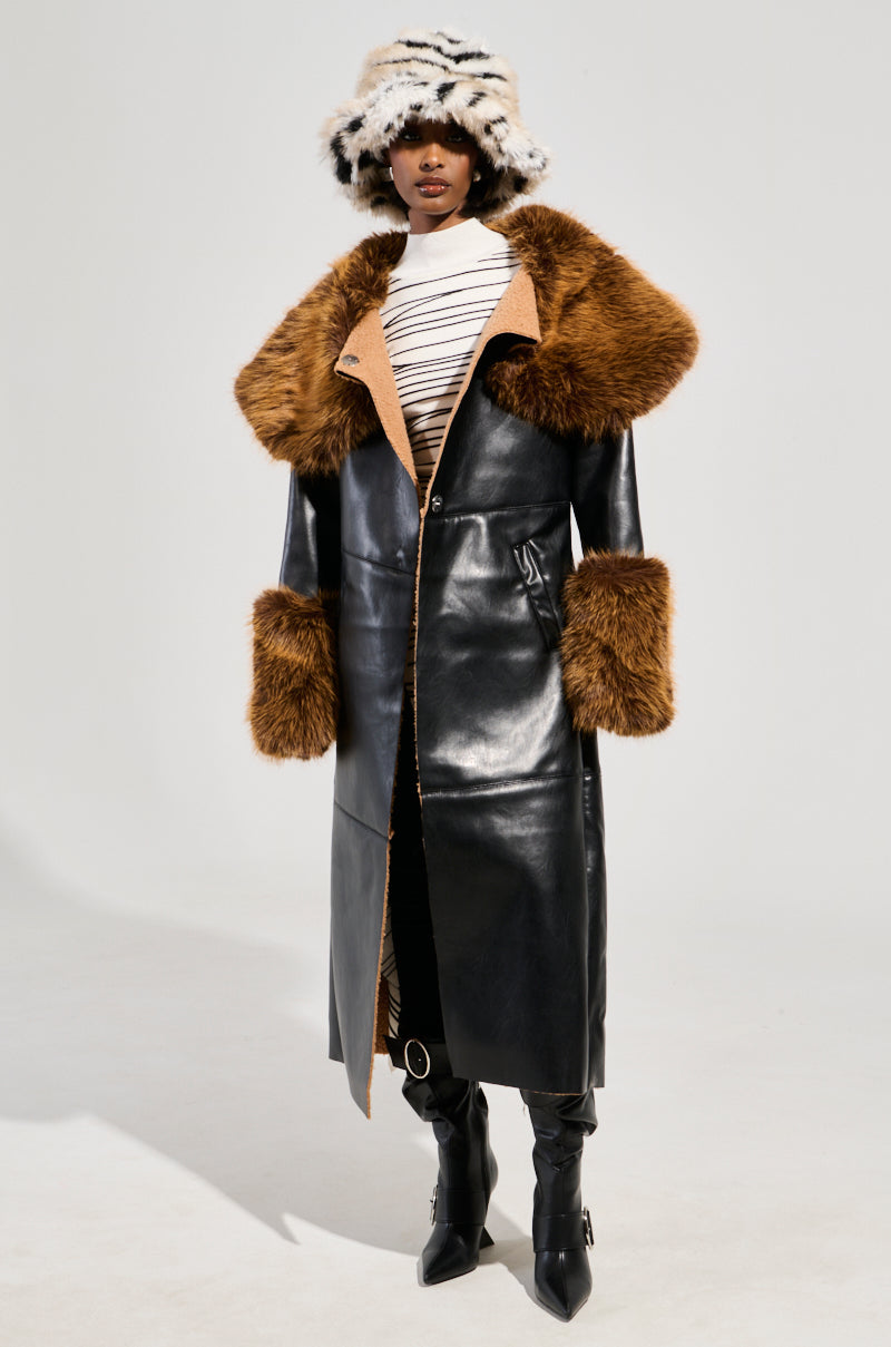 The individual is dressed in the SIREN FUR TRIM TRENCH COAT, a long black faux leather trench coat with large brown faux fur trim on the collar and cuffs. They pair it with a white and black striped top, black knee-high boots, and a black and white fuzzy hat. The background is plain gray.