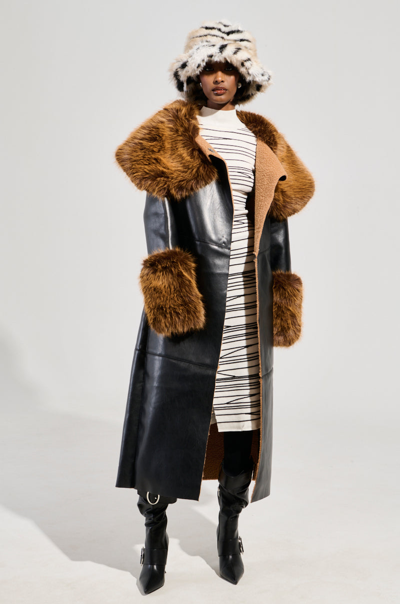 A person wearing the SIREN FUR TRIM TRENCH COAT, a long black faux leather trench coat with large brown faux fur trim cuffs and collar, paired with a white striped dress, black heeled boots, and a fluffy black and white hat stands against a plain background.