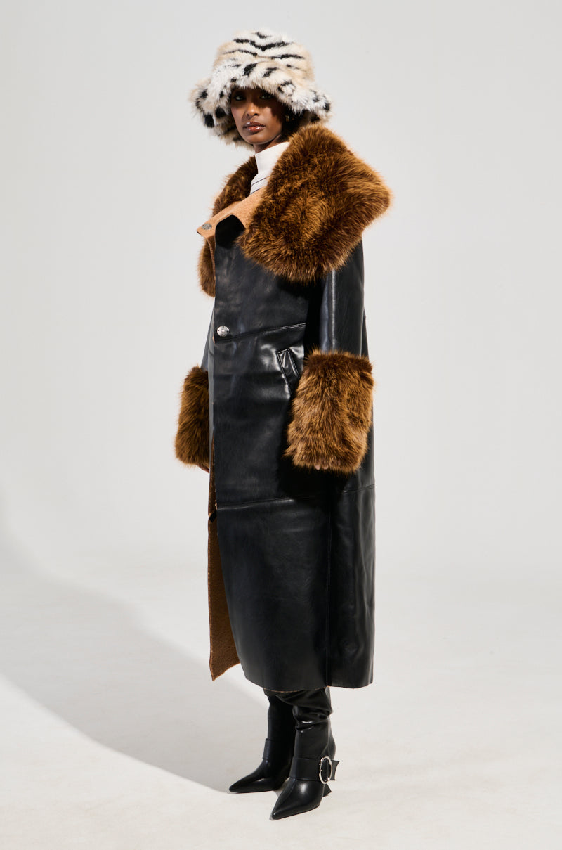 A person is wearing the SIREN FUR TRIM TRENCH COAT, a long black faux leather trench coat with a brown faux fur trim collar and cuffs. They pair it with a white and black striped fur hat and black pointy boots, standing against a plain background.