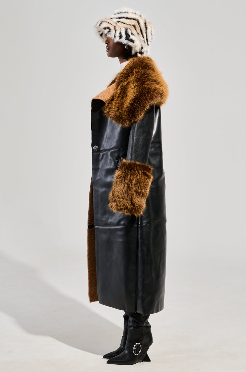 A person wearing the SIREN FUR TRIM TRENCH COAT with brown faux fur trim on the collar and cuffs, a zebra-patterned hat, and black boots with metal accents, standing in profile against a plain background.