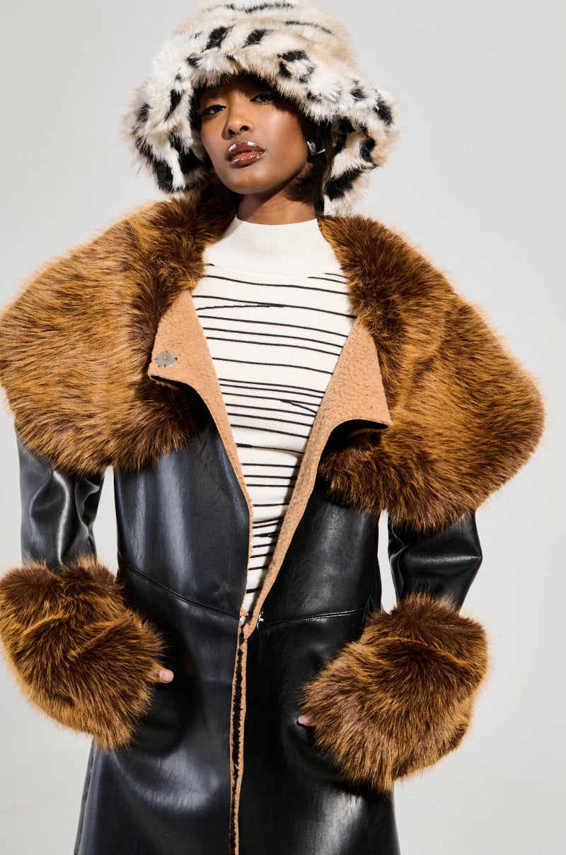 A person wearing a stylish outfit featuring the SIREN FUR TRIM TRENCH COAT in black faux leather with a brown faux fur trim over a striped sweater, accessorized with a fluffy leopard-print hat. The background is plain gray.