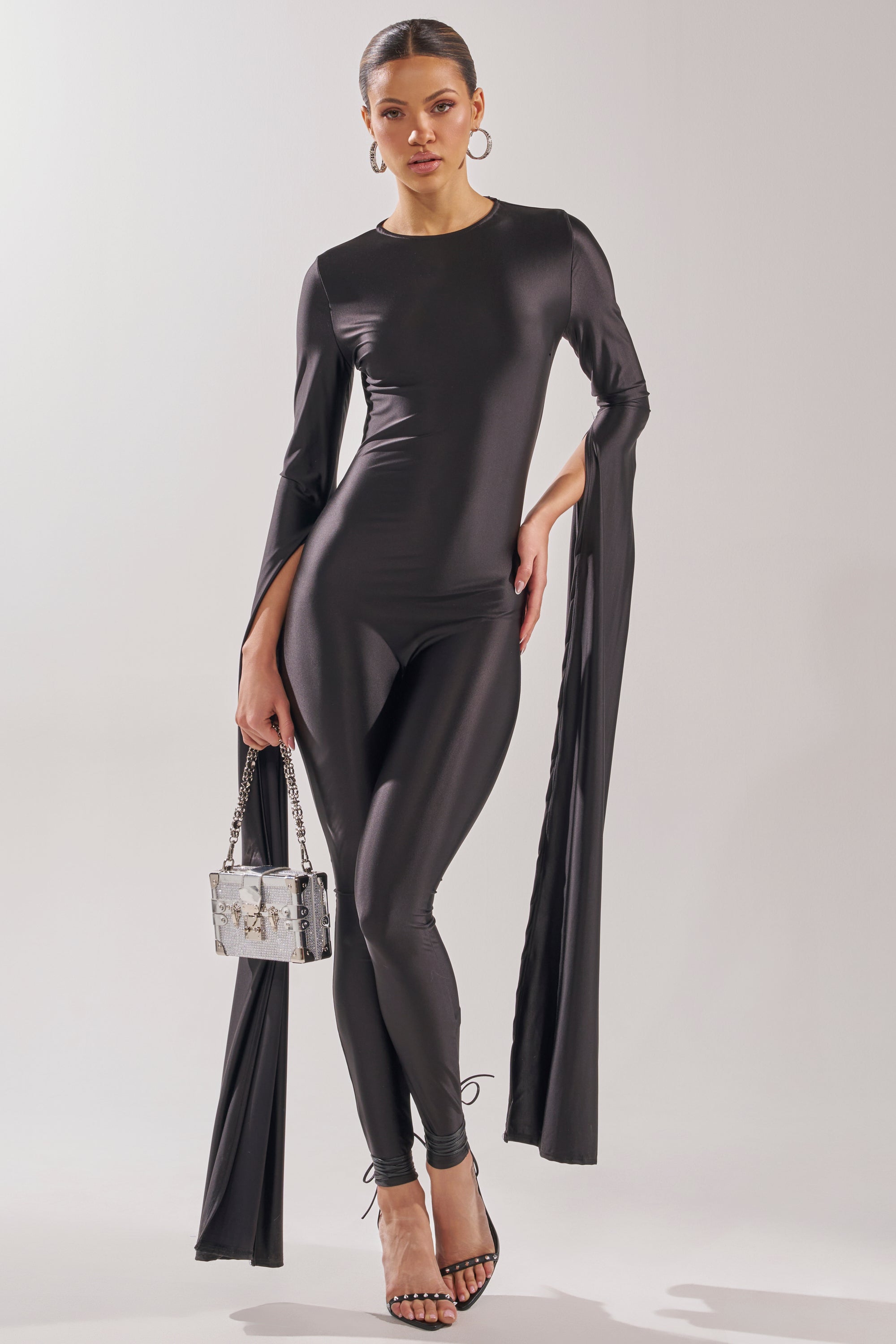 A woman models the CATWALK QUEEN JUMPSUIT—a sleek, black, skinny-leg style with long draping sleeves. She pairs it with strappy black heels, a silver handbag, hoop earrings, and a pulled-back hairstyle. 0