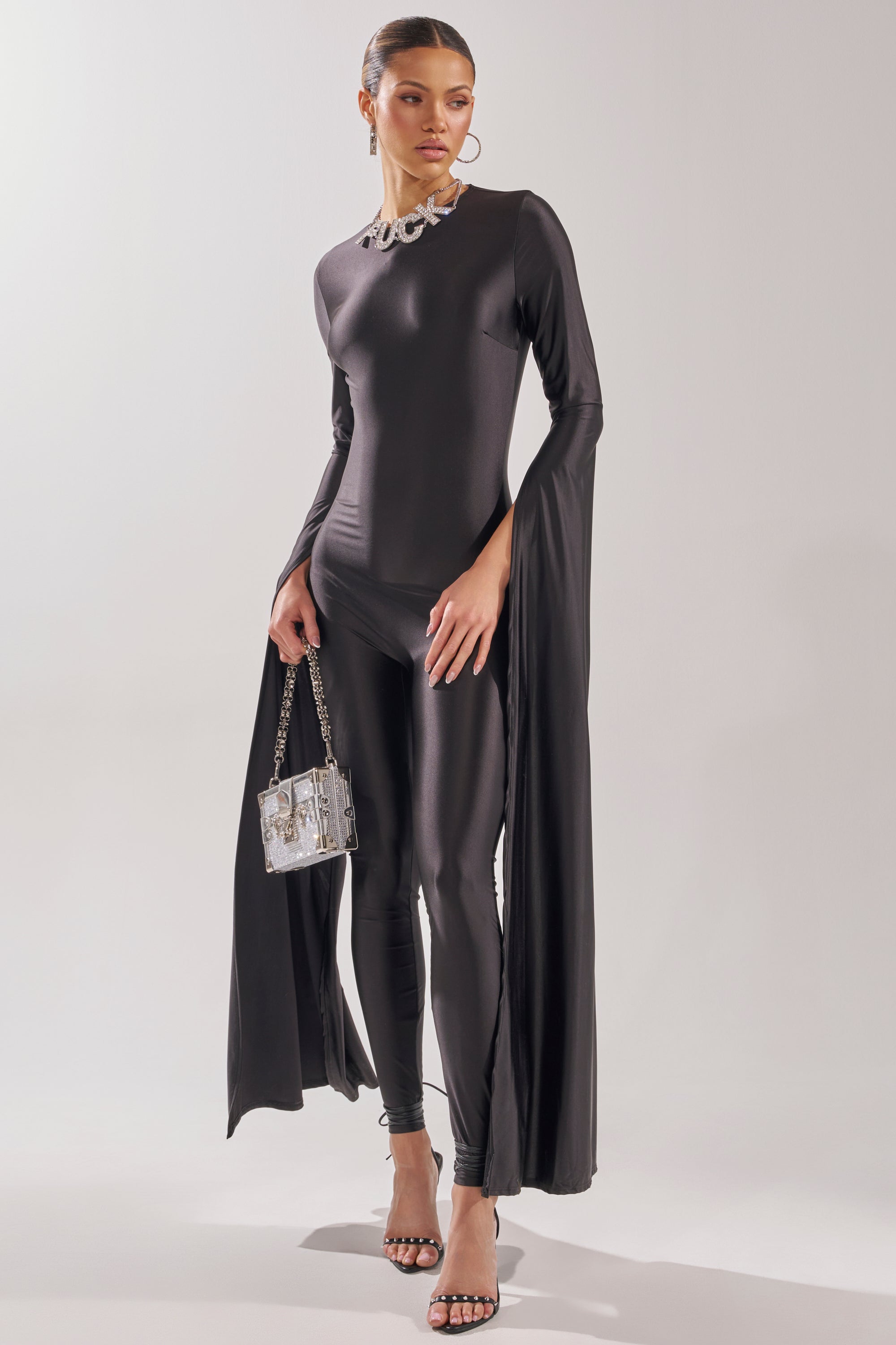 A woman stands against a plain light background wearing the CATWALK QUEEN JUMPSUIT, featuring dramatic cape sleeves and a skinny-leg fit, styled with strappy high heels, a statement necklace, hoop earrings, and a small shiny handbag. 1