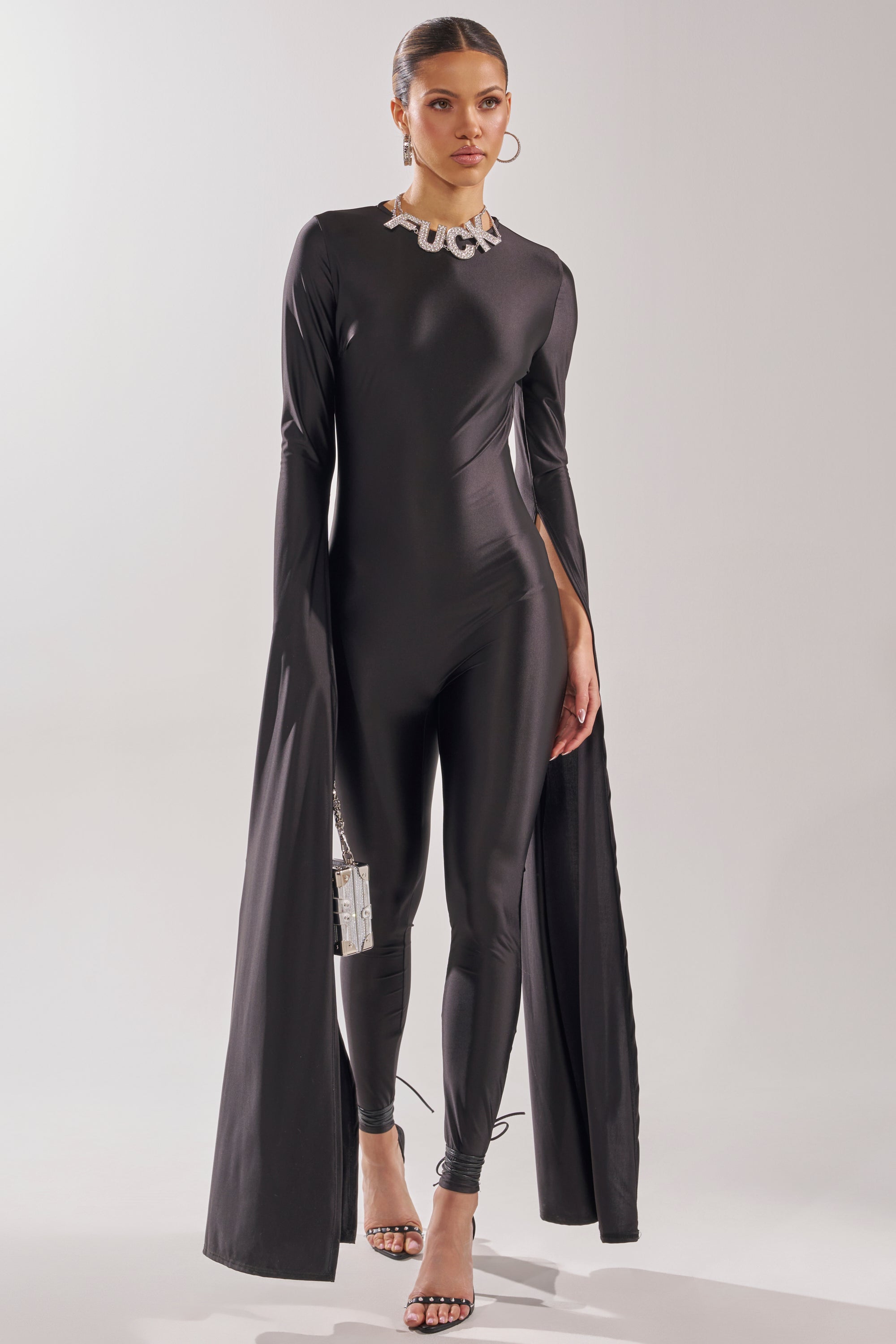 A woman models the CATWALK QUEEN JUMPSUIT—a sleek black design with floor-length draped sleeves and skinny legs. She accessorizes with a small purse, hoop earrings, open-toe heels, and a bold “FUCK” statement necklace. 2