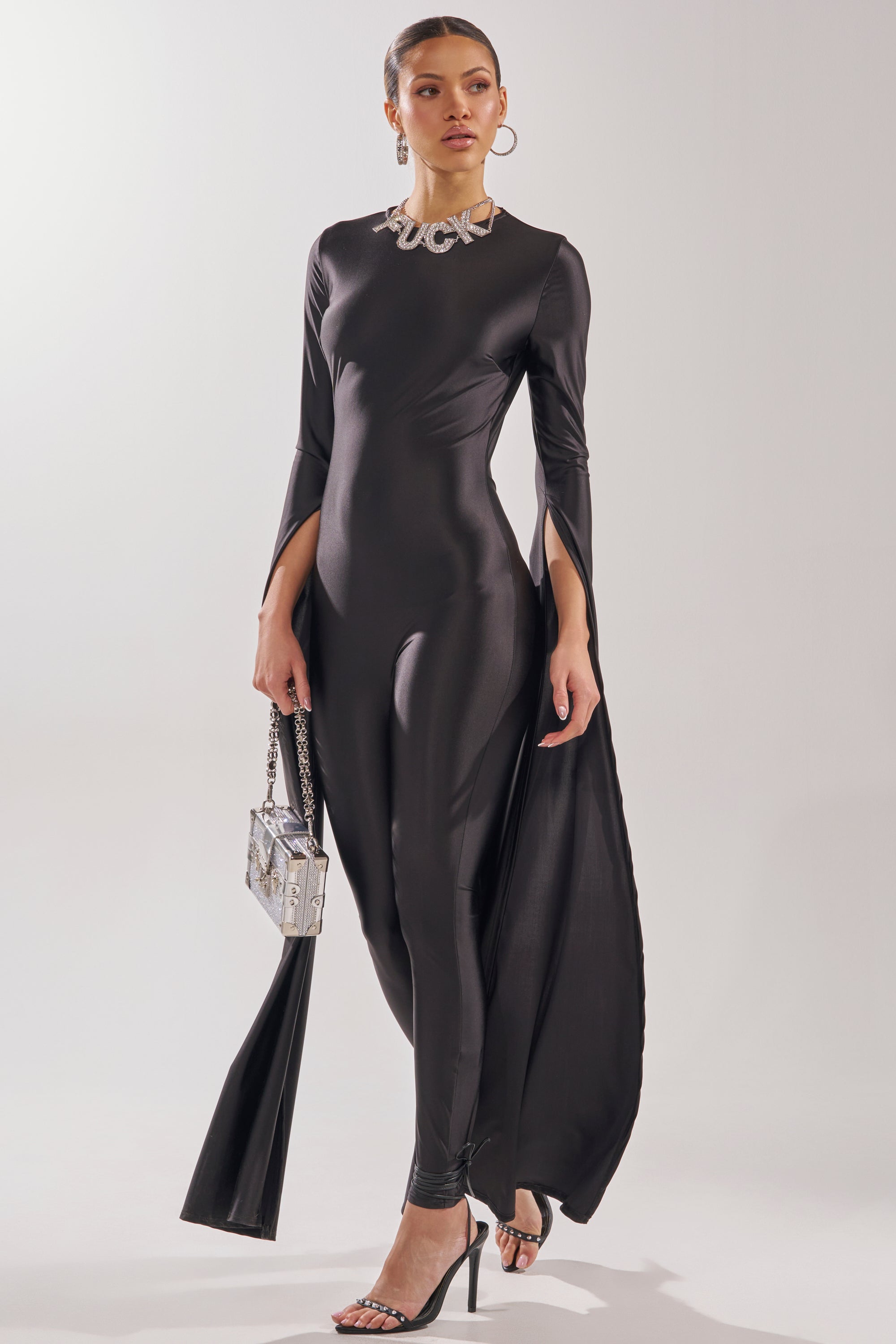 A woman poses confidently against a plain background in the CATWALK QUEEN JUMPSUIT—a sleek, long-sleeve black piece with draped panels and an open back—styled with black heels, hoop earrings, a chunky silver necklace, and a metallic handbag. 3