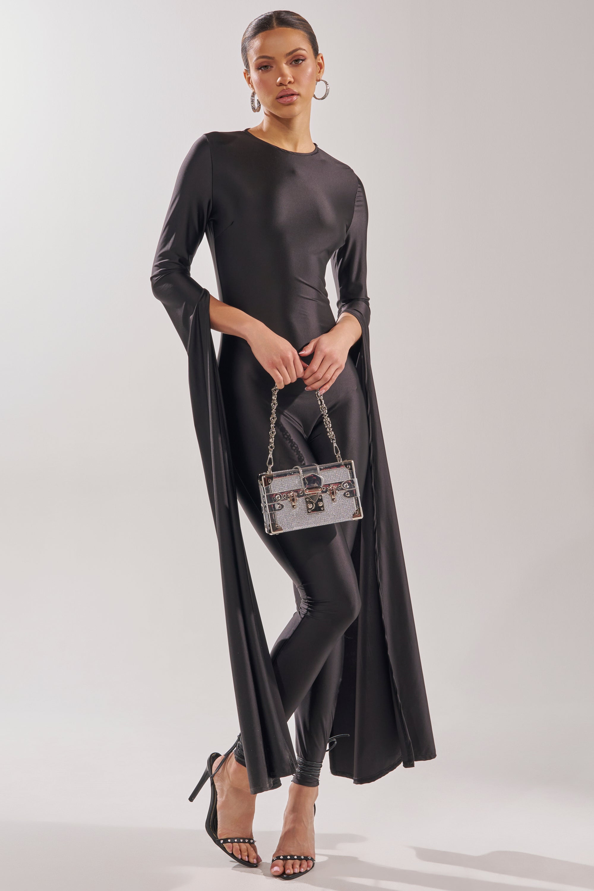 A woman exudes confidence in the CATWALK QUEEN JUMPSUIT, featuring skinny legs and cape sleeves. She pairs it with hoop earrings, strappy heels, a silver handbag, and a sleek pulled-back hairstyle against a light backdrop. 4