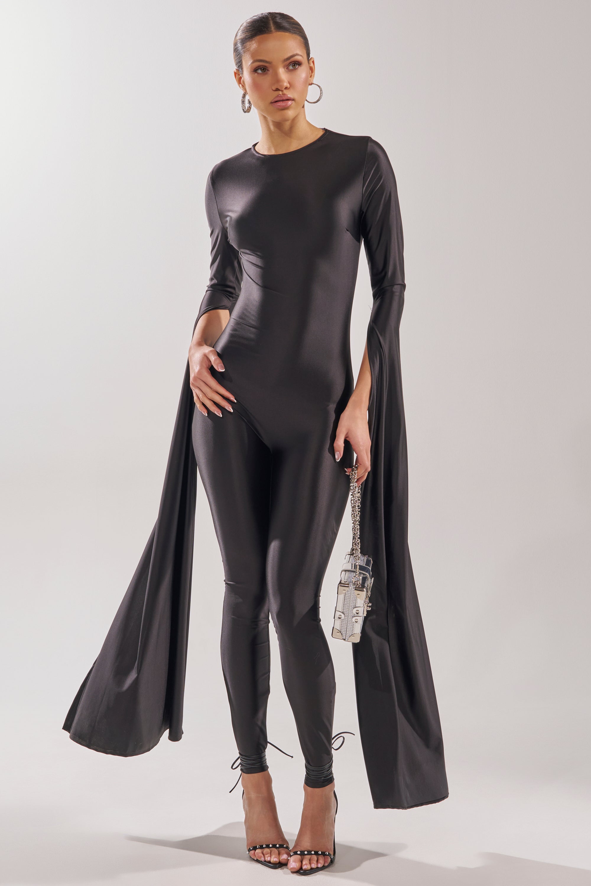A woman models the CATWALK QUEEN JUMPSUIT, featuring dramatic cape-like sleeve panels. She accessorizes with a small silver handbag, hoop earrings, and strappy heels, posing confidently against a simple backdrop. 5