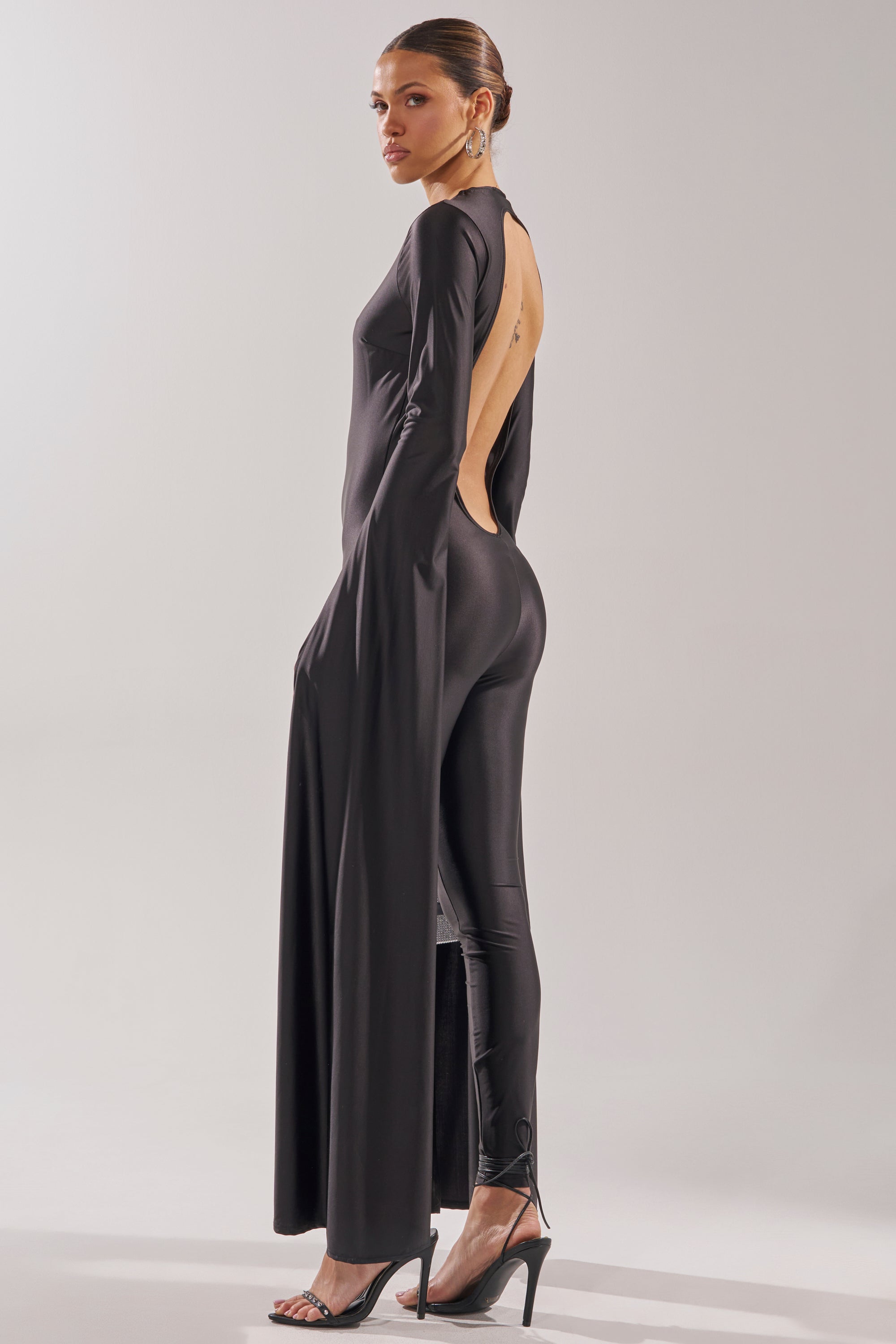 A woman poses in profile wearing the CATWALK QUEEN JUMPSUIT, featuring an open back and flowing train-like panels. She pairs the sleek black jumpsuit with high-heeled sandals and has her hair pulled back against a neutral background. 6