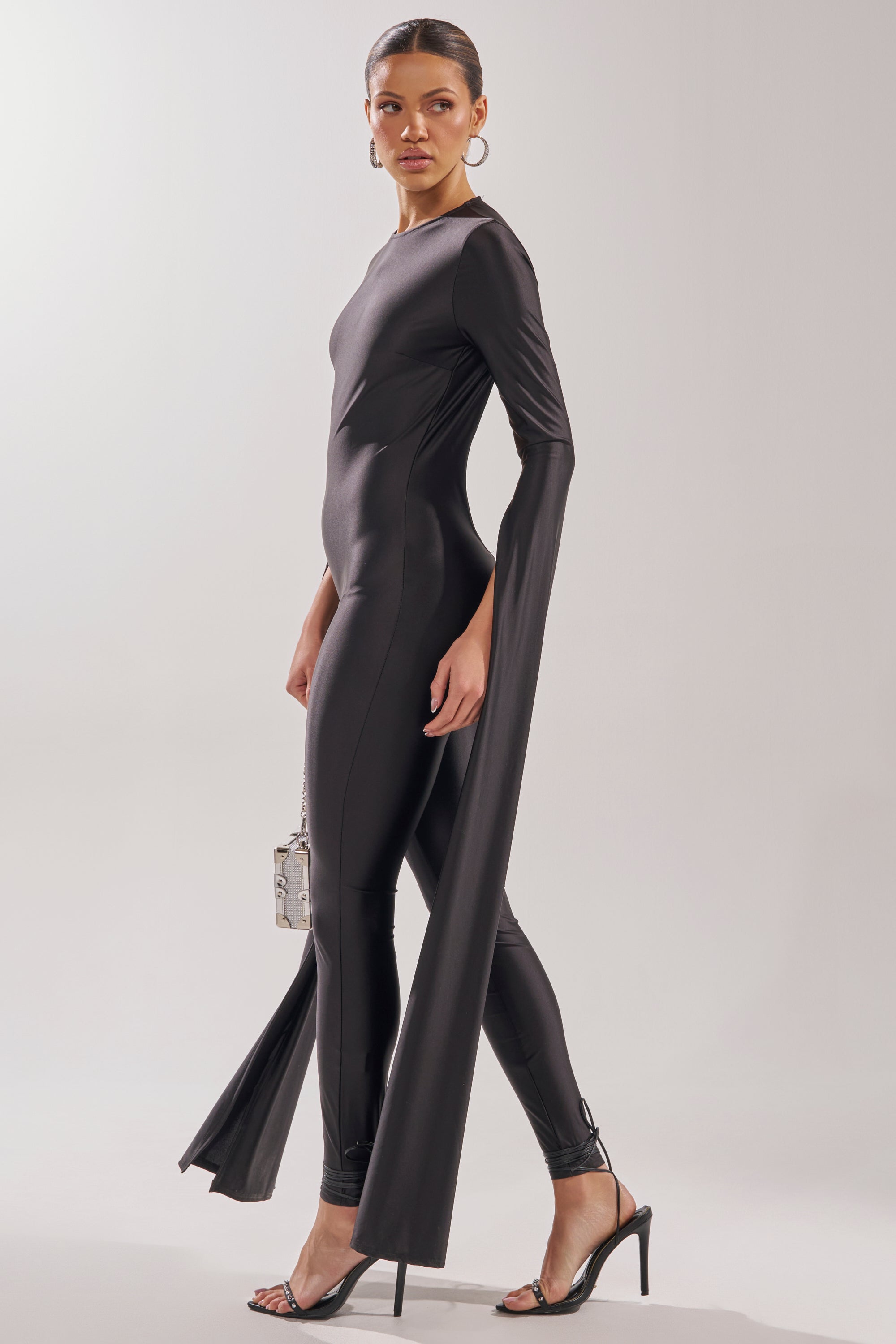 A woman stands against a plain light background wearing the CATWALK QUEEN JUMPSUIT—a sleek black skinny-leg jumpsuit with dramatic flared long sleeves—styled with black heeled sandals, gold hoop earrings, and a small metallic handbag. 7