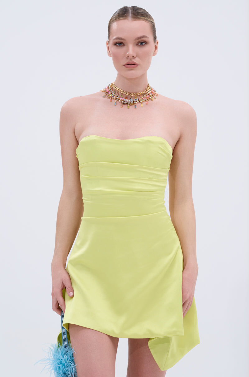 A woman wears the LET'S ESCAPE SATIN MINI DRESS, a strapless light yellow mini with an asymmetrical hem. She styles it with a colorful beaded choker and holds a light blue feathered accessory against a plain white background.