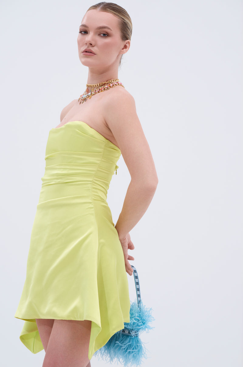 A woman stands against a plain background, wearing the LET'S ESCAPE SATIN MINI DRESS—a strapless, light yellow mini dress—layered necklaces, and holding a small blue feathered purse behind her back.