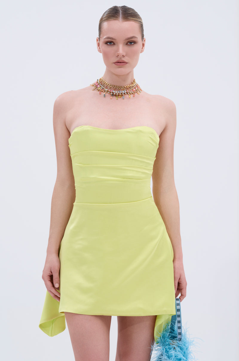 A woman in the LET'S ESCAPE SATIN MINI DRESS, a strapless light yellow mini dress, stands against a plain backdrop. She wears a colorful beaded choker and holds a blue feathery accessory in her right hand.
