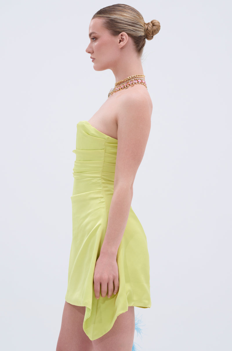 A woman in the LET'S ESCAPE SATIN MINI DRESS in light yellow stands in profile against a plain background, accessorized with a gold necklace and her hair styled in a neat bun.