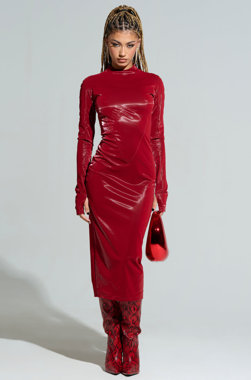 A person with long braids wears the LIKE LIQUID FAUX LEATHER MIDI DRESS in a sleek, red design, paired with matching knee-high boots. They hold a shiny red handbag and stand against a plain background.