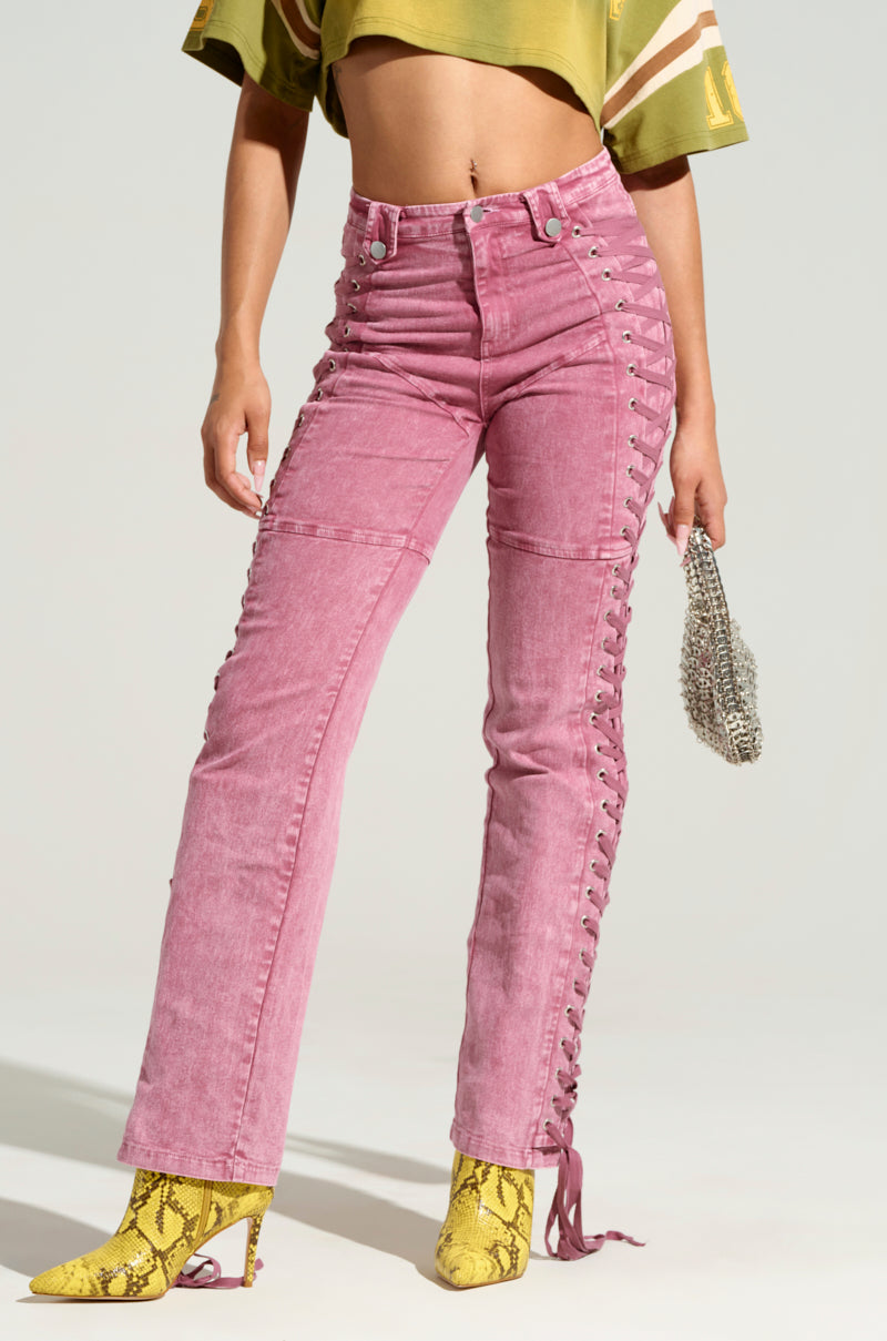 A person is dressed in pink STAND OUT LACED UP JEANS, paired with a cropped green top and yellow snake-print heeled boots. They are holding a small, sparkly silver bag, with the focus on their legs and waist against a neutral light background.