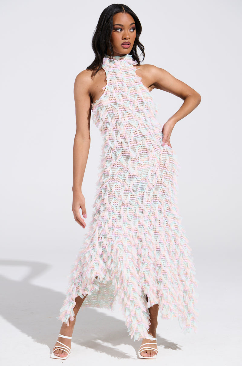 A woman poses confidently in the IT FACTOR MESH MAXI DRESS IN PINK, featuring a pastel and fringe-like texture. She completes her look with white heeled sandals, standing against a plain white background with her hair styled down.