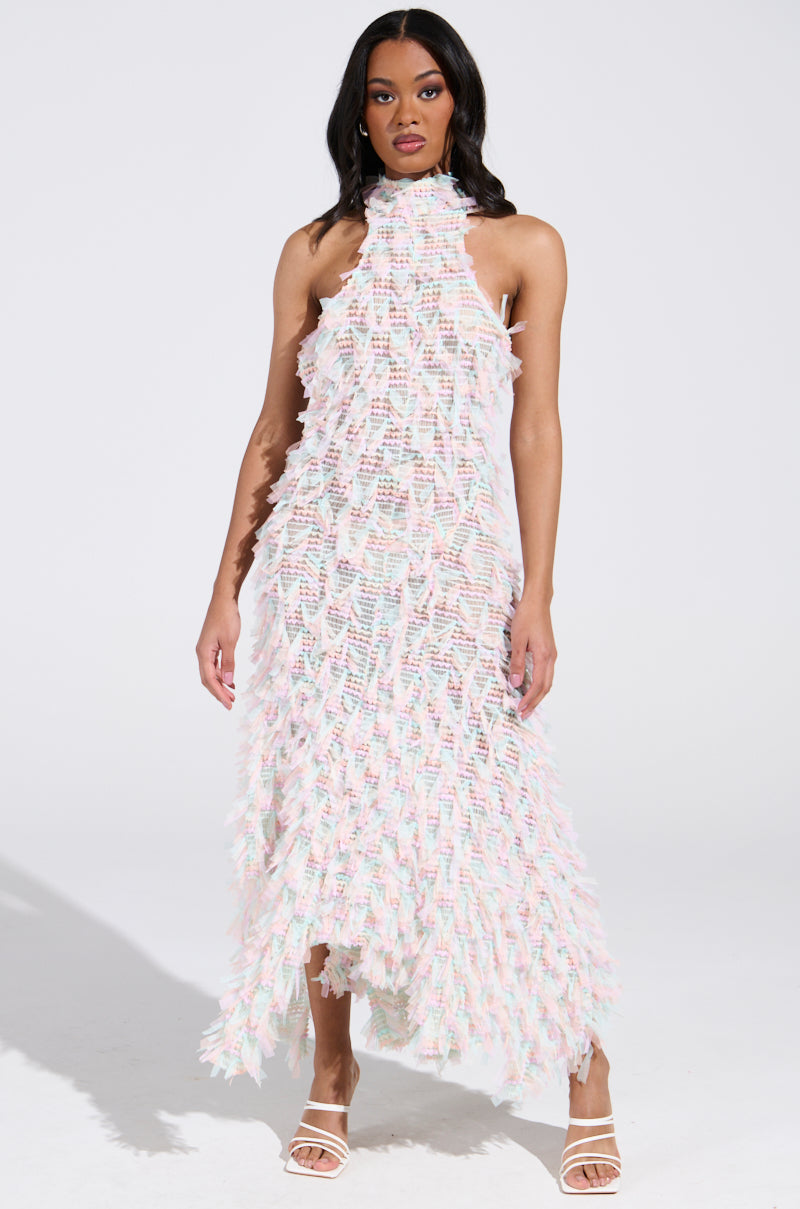 The person poses against a plain white background wearing the IT FACTOR MESH MAXI DRESS IN PINK with a sleeveless, fit-and-flare silhouette showcasing a textured, feathery pattern in pastel shades, complemented by white strappy high heels.