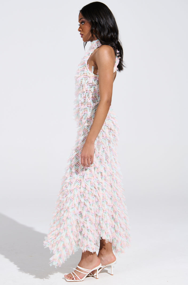 Against a white backdrop, a woman with long dark hair models the "It Factor Mesh Maxi Dress in Pink," a sleeveless pastel gown featuring a textured finish, fringes, and a fit-and-flare design. Her look is completed with white heeled sandals, exuding elegance.