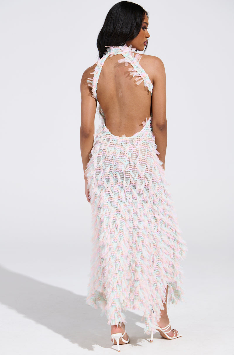 Wearing the elegant IT FACTOR MESH MAXI DRESS IN PINK, a person showcases their style. The sleeveless, open-back dress features multicolored feather-like textures and flows to mid-calf. Their long dark hair complements white heeled sandals as they stand on a white surface.