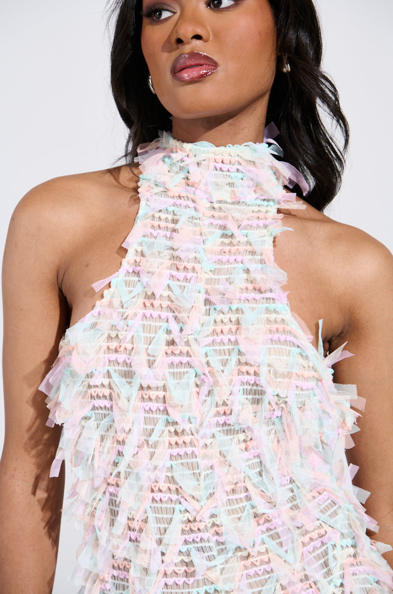 Wearing the IT FACTOR MESH MAXI DRESS IN PINK, featuring pastel hues, textured geometric patterns, and a fit-and-flare silhouette with fringe layers and a high neckline, she captivates with her cascading dark hair, glossy makeup, and stylish hoop earrings.