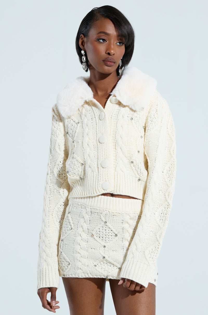 Against a bright backdrop, an individual is dressed in a cream knit set that features a cozy faux fur collar. The ensemble includes the cropped Snow Bunny Sweater, complemented by a matching skirt that showcases an intricate textured design. With shoulder-length hair and long, dangling earrings, the look is both chic and stylish. 0
