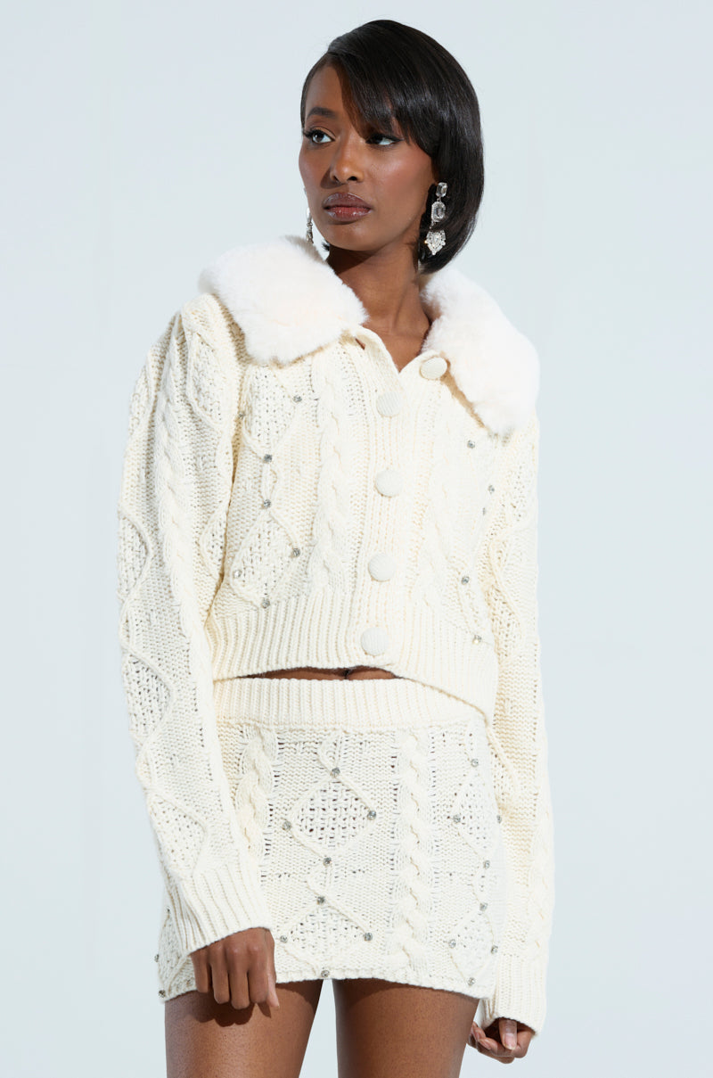 A person wearing the SNOW BUNNY SWEATER, a cream-colored knit set adorned with a fluffy faux fur collar and button detailing. The outfit showcases an elegant diamond pattern. They complement their look with long earrings and are gazing thoughtfully to the side against a plain background. 1