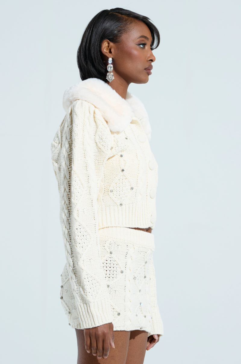 A woman in profile sports a knitted cream-colored SNOW BUNNY SWEATER adorned with rhinestone details and a faux fur collar. Her outfit includes a long-sleeved top paired with a skirt. Complementing her short, sleek hair, she adds dangling earrings for accessories, set against a light, neutral background. 2