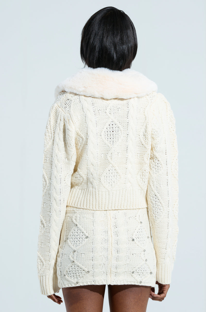 A person with short black hair stands facing away, dressed in the cream-colored SNOW BUNNY SWEATER dress, featuring a textured diamond pattern and rhinestone embellishments. The long sleeves beautifully complement the faux fur collar, all set against a neutral light gray background. 3