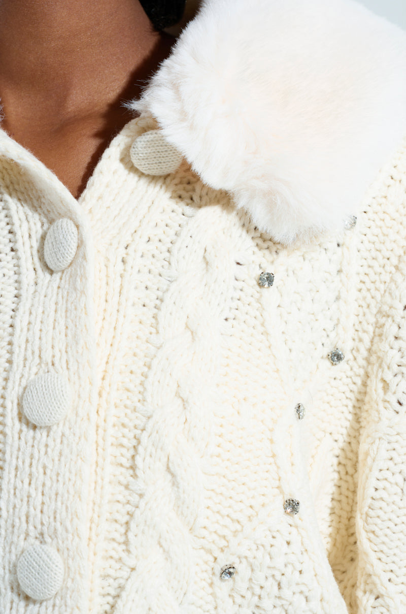 A detailed view of the SNOW BUNNY SWEATER reveals its cream-colored knit design with prominent buttons. It boasts a cable knit pattern and is elegantly finished with a plush faux fur collar. The sweater is beautifully accented with small, sparkling embellishments, possibly rhinestones or sequins. 6