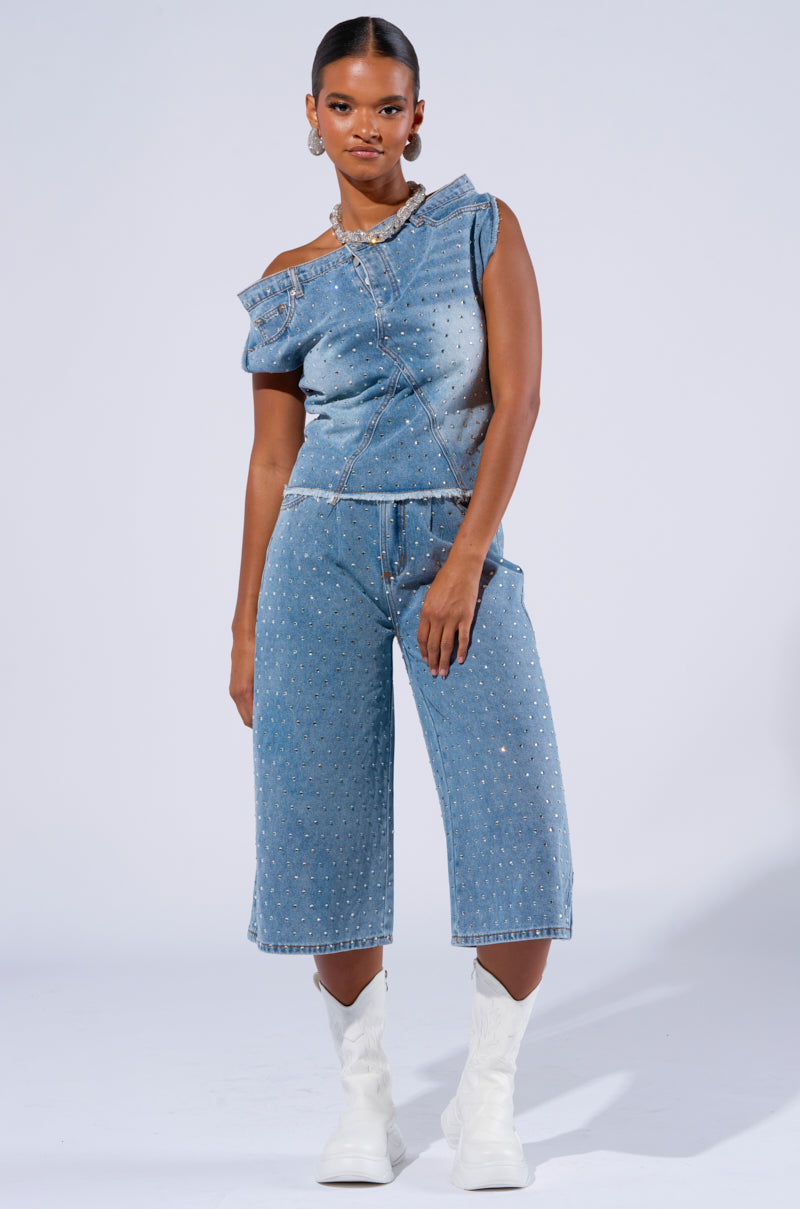 A woman is standing against a plain background, wearing a stylish off-shoulder denim top paired with DENIM DIVA SHORTS, which are beautifully adorned with AB rhinestone embellishments. She completes the look with white ankle boots and a statement necklace, looking poised and fashionable.