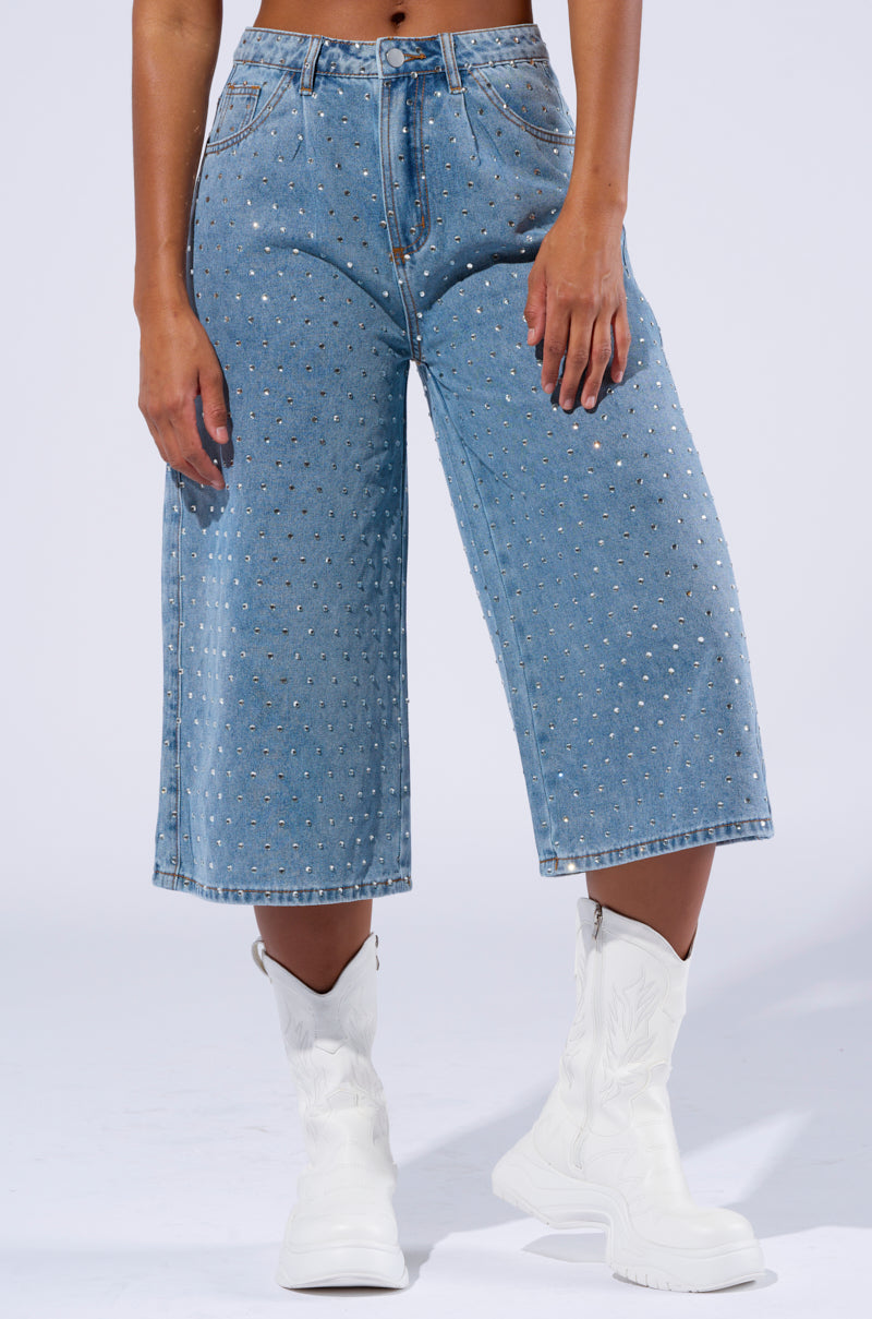 A person is modeling the DENIM DIVA SHORT, high rise waist knee-length light blue denim pants adorned with small AB rhinestone embellishments, paired with white cowboy boots. The background is plain light gray. The image focuses on the lower half of the person's body, displaying the outfit.