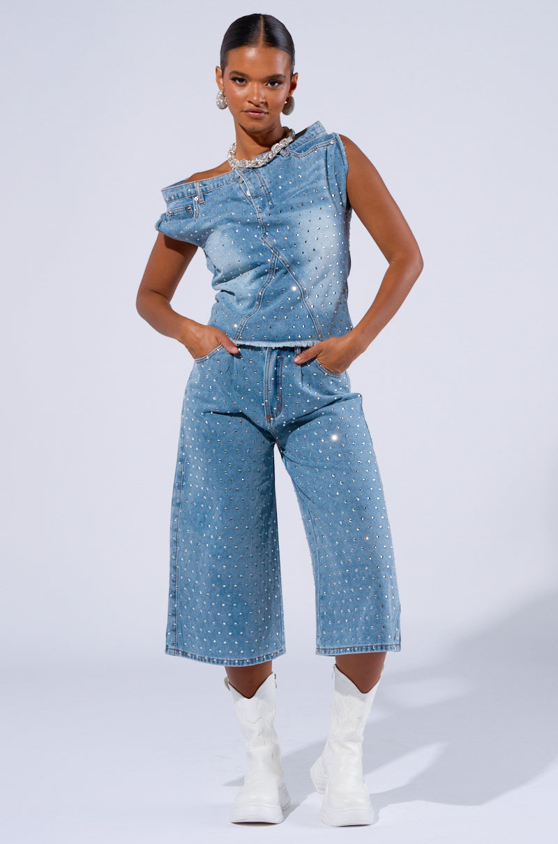 A model confidently poses in a matching denim outfit adorned with AB rhinestone embellishments. She wears a distinctive sleeveless denim top featuring an asymmetrical neckline, paired with high-waisted, knee-length DENIM DIVA SHORTS. The look is completed with white ankle boots and statement earrings.