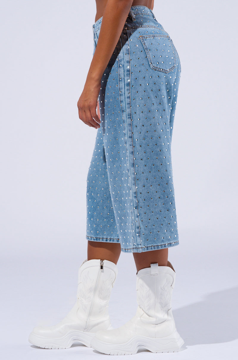 Side view of a person wearing the DENIM DIVA SHORT from AKIRA Label, featuring a high-rise waist and rhinestone embellishments. They are also sporting white, chunky, calf-high boots with intricate stitching. The image focuses on the lower half of the body against a plain background.