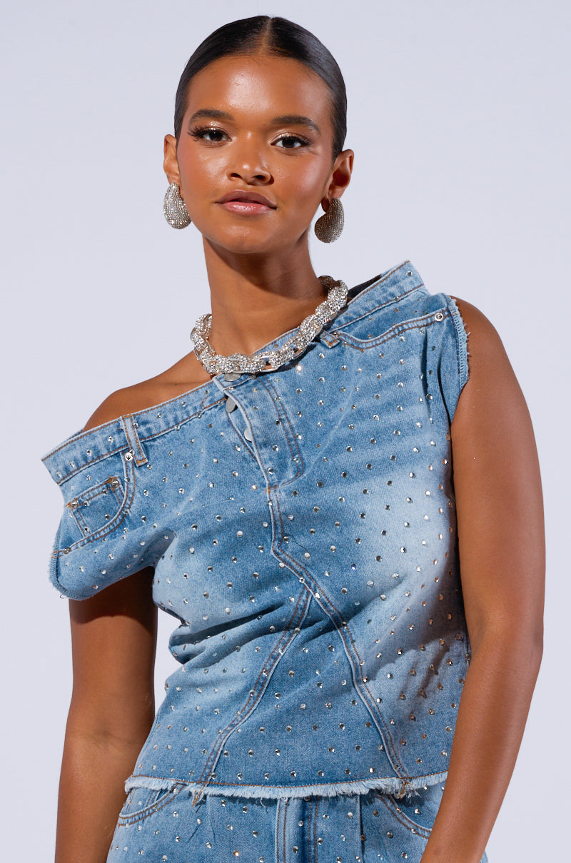 A woman stands against a plain background, embodying the Denim Diva look with the DENIM DIVA OFF SHOULDER TOP, featuring asymmetrical denim design adorned with AB rhinestone embellishments. She accessorizes with large silver hoop earrings and a statement necklace. Her hair is pulled back, and she has a confident expression.