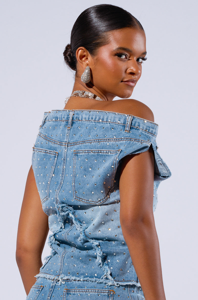A woman with smooth, dark skin and her hair styled in a sleek bun poses with a confident expression. She is the epitome of a Denim Diva, sporting the DENIM DIVA OFF SHOULDER TOP adorned with AB rhinestone embellishments. Her sparkling earrings and necklace add to the glamour against the plain, light-colored background.
