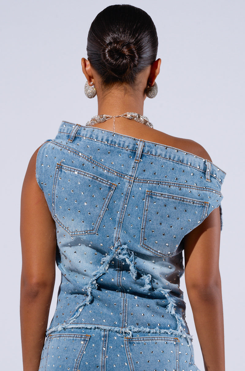 From behind, a person with slicked-back hair wears the DENIM DIVA OFF SHOULDER TOP adorned with small silver studs and distressed elements. The top drapes off one shoulder, revealing a silver chain necklace and large, circular silver earrings accented with AB rhinestone embellishments.