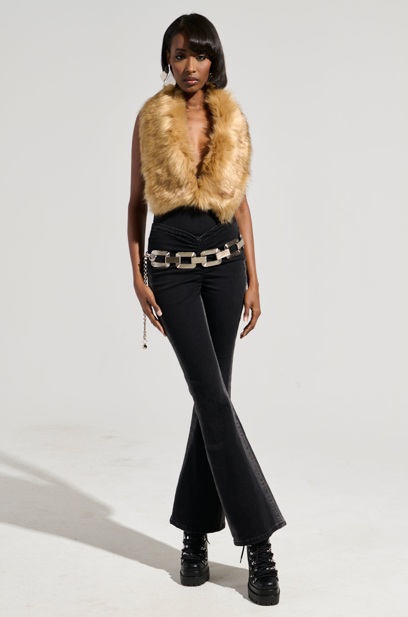A person stands confidently against a plain background, showcasing a sleeveless fur vest with faux fur detailing, paired with black flared pants and platform shoes. They accessorize with an edgy chain belt and embody the chic flair reminiscent of the FEELS GOOD BODYSUIT, exuding a stylish and poised presence.