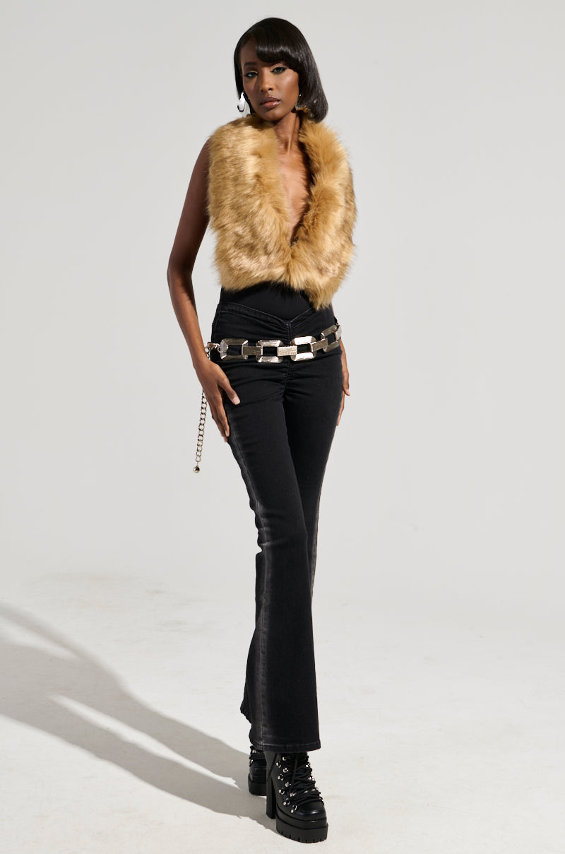 A person stands confidently against a plain background, wearing the FEELS GOOD BODYSUIT with faux fur detailing, paired with black high-waisted pants featuring a chain belt and black platform shoes. Their short, sleek hair complements their pose, with hands on their hips.