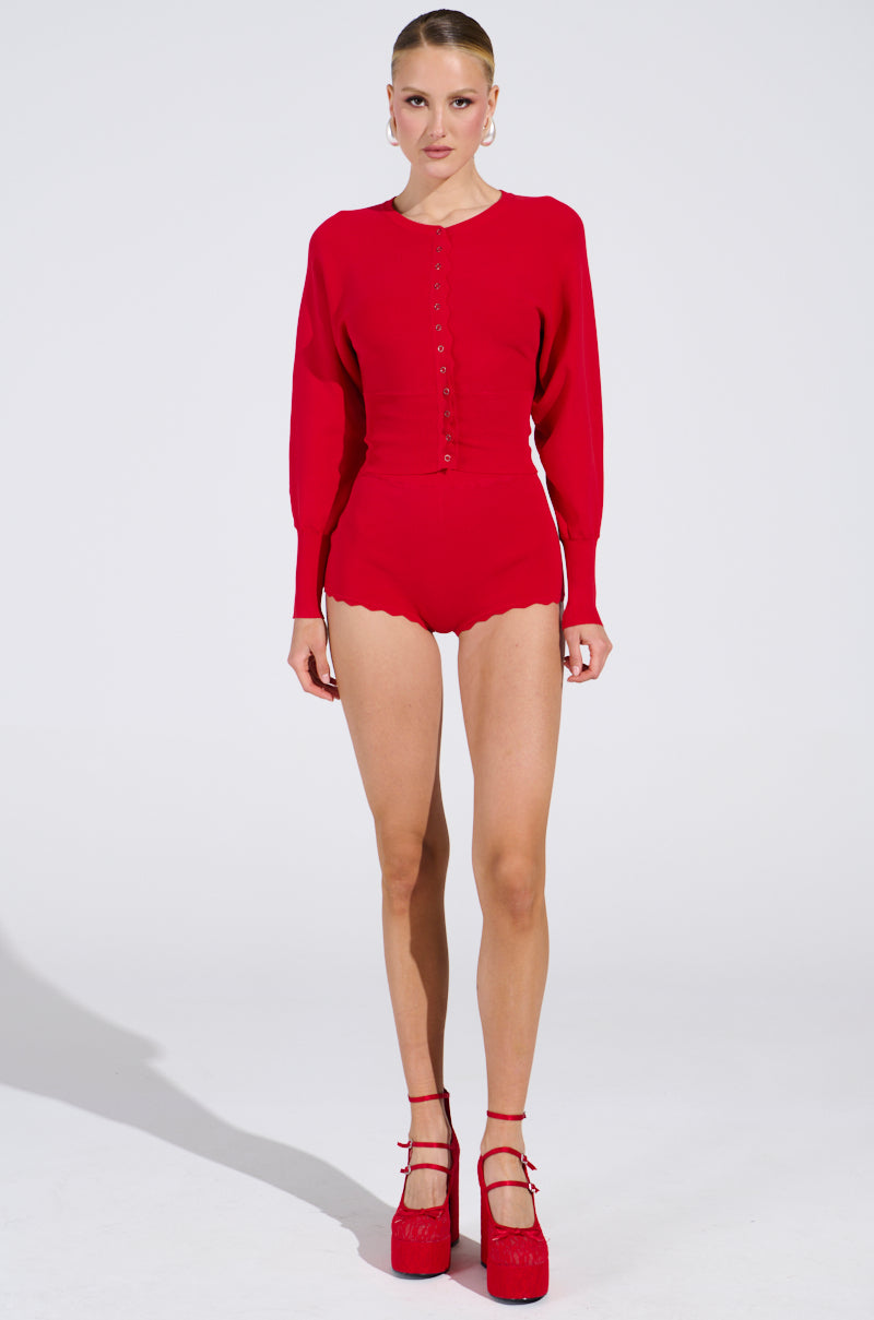 A person is wearing a bright red, long-sleeve top and the BRING ME LUCK HOT SHORT—high-waisted with a curve-hugging fit. They are standing against a white backdrop, sporting matching red heels and hoop earrings.