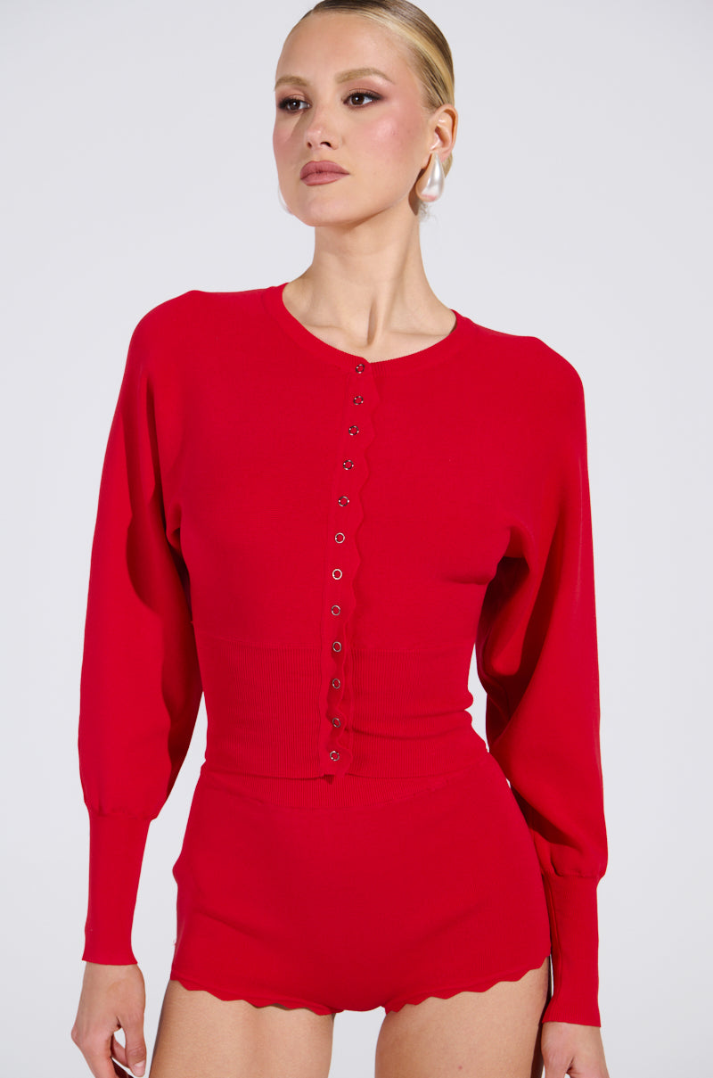 A model displays a red, semi-stretchy knit top with button details and matching high-waisted bottoms. The scalloped edges on the sleeves and shorts evoke the BRING ME LUCK CARDIGAN. The cropped silhouette enhances her pose against a plain white background.