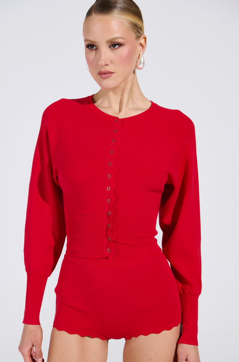 Dressed in a vibrant red, semi-stretchy knit bodysuit with high-waisted shorts, scalloped edges, and fitted cuffs reminiscent of the sleek BRING ME LUCK CARDIGAN. The background is plain and their hair is neatly pulled back.