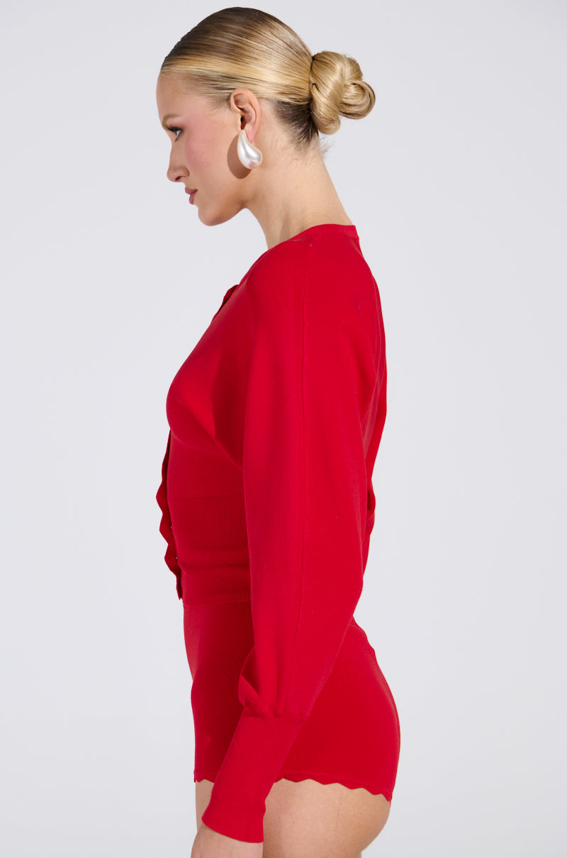 A person with blonde hair in a bun wears a red, form-fitting outfit with scalloped edges and a cropped look. They have large round white earrings and face left against a neutral backdrop, echoing the charm of the BRING ME LUCK CARDIGAN.