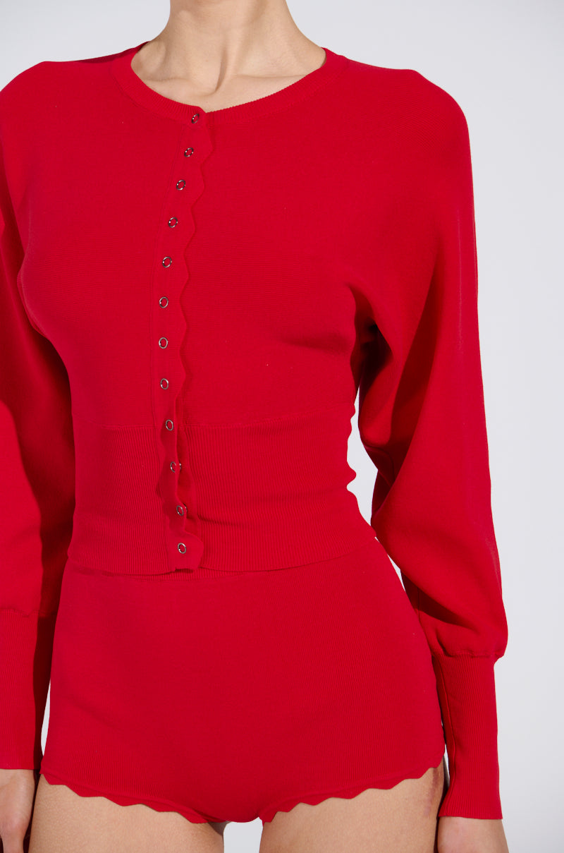 The image shows a person in a vibrant red, semi-stretchy knit outfit with a snug, high-waisted fit. The ribbed fabric has scalloped edges and button details similar to the BRING ME LUCK CARDIGAN. The stylish design stands out against the plain white background.