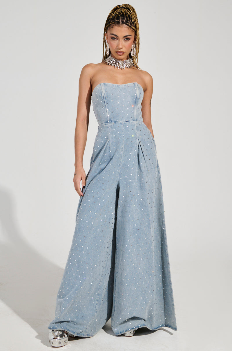 Dressed in the "CAN'T OUTSHINE ME" denim and rhinestone jumpsuit, a person exudes confidence. The strapless design features dazzling embellishments in a light wash hue. Complementing their look is a beaded choker and intricately braided hair as they pose stylishly against a plain background.